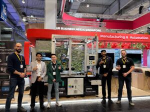 Five team members from Augmentus Robotics and Servind Automation giving thumbs up at Hannover Messe trade show, standing in front of an automated sanding cell featuring a Universal Robots cobot arm and Mirka abrasive system, inside the Singapore pavilion's Manufacturing & Automation zone.